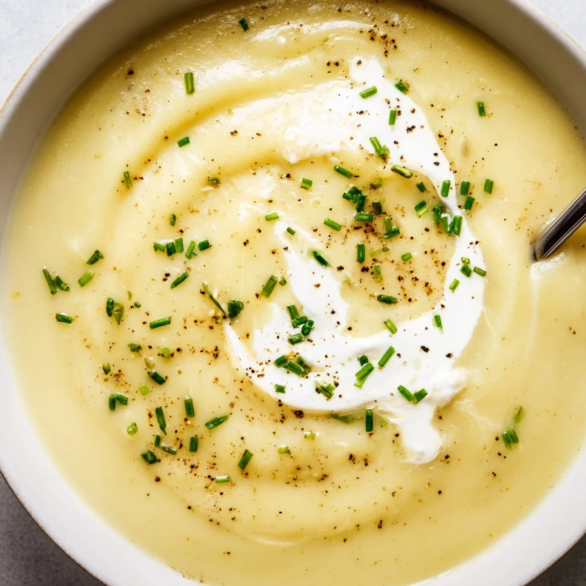 A spoon dips into velvety Creamy Leek Soup with Potatoes, showcasing the smooth, pale green puree alongside crusty artisan bread.