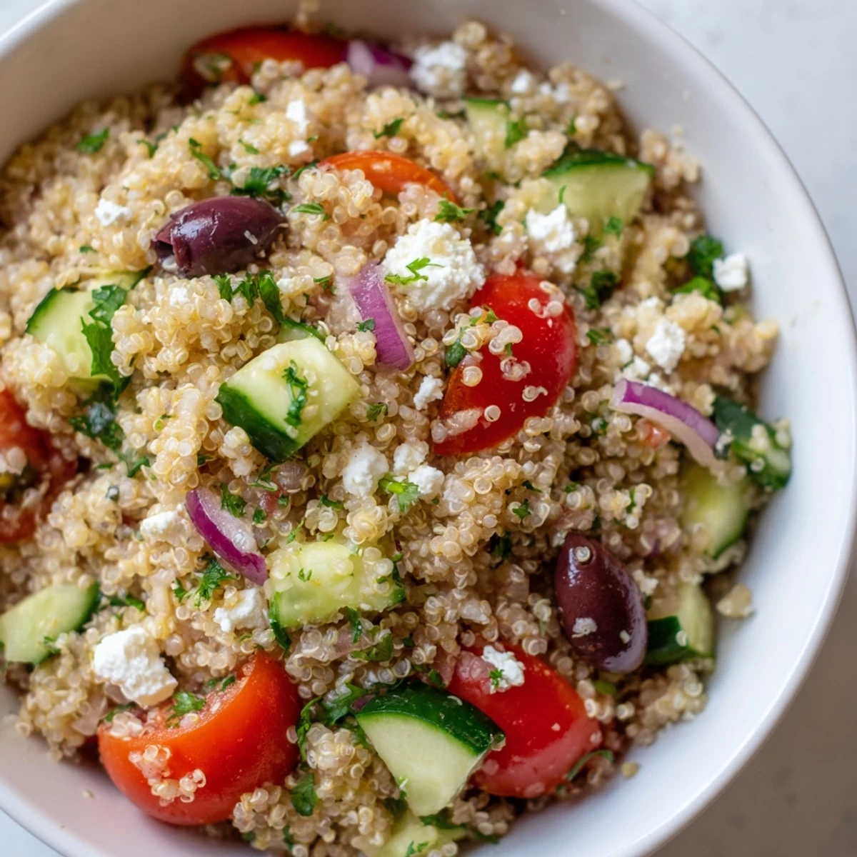 A close-up shows vibrant red tomatoes, crisp green cucumbers, and purple olives mixed into fluffy quinoa with feta cheese.