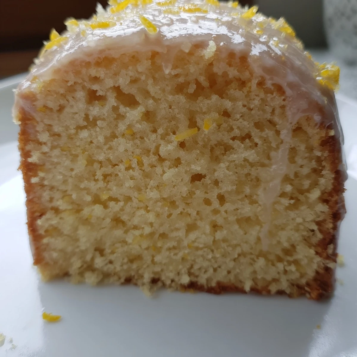 A slice of Lemon Drizzle Cake is plated with fresh raspberries and a dollop of whipped cream, highlighting the moist crumb.
