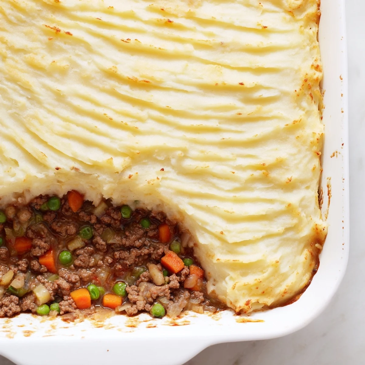 A golden-brown Shepherds Pie with ground beef filling peeking through creamy, fork-ridged mashed potatoes, served in a rustic baking dish.  