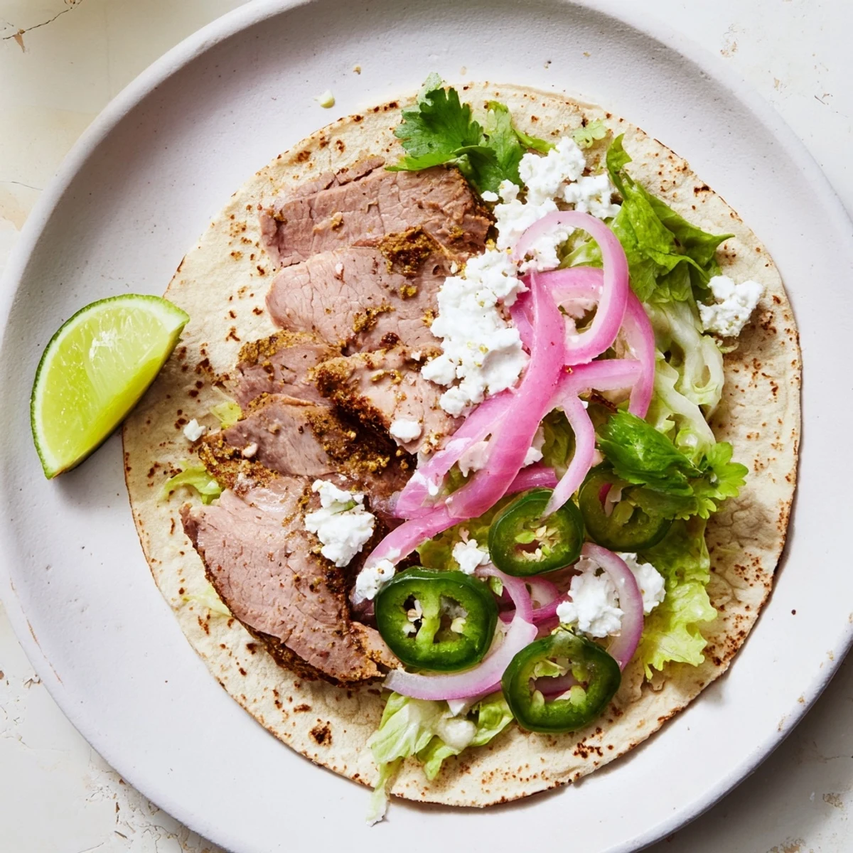 Shiny, slow-roasted cumin crusted leg of lamb with tacos on a plate, drizzled with creamy yogurt-lime sauce and garnished with fresh jalapeño slices.