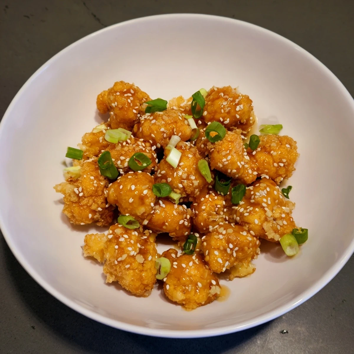 Golden-brown, crispy Sticky Honey Garlic Cauliflower florets glistening with sticky sauce and sprinkled with sesame seeds and green onions on a white plate.