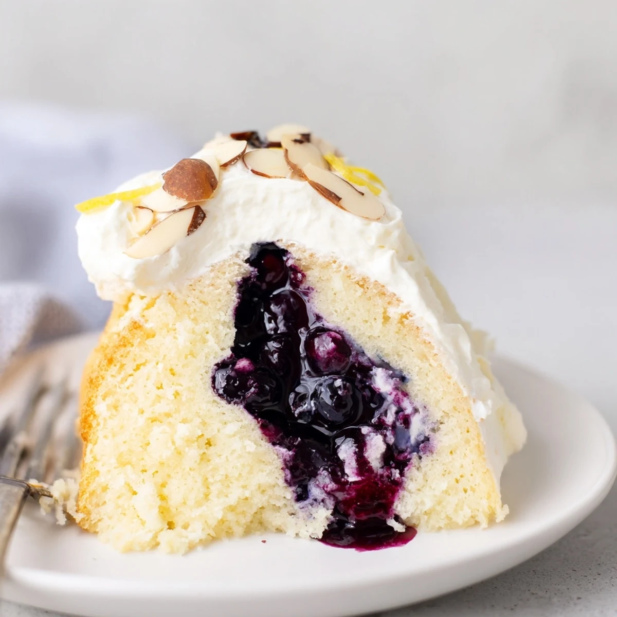 Sliced Lemon Blueberry Poke Cake With Almond Topping on white plate, glossy blueberry pockets.
