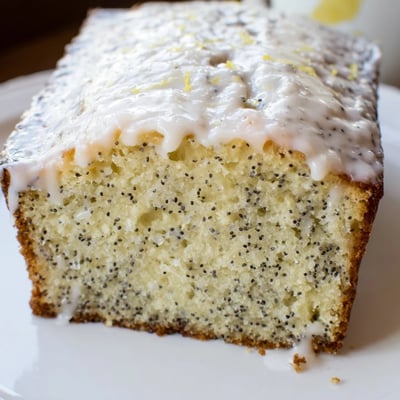 Perfectly moist Lemon Poppy Seed Loaf slice on a white plate, with a cup of coffee and fresh lemon wedges for a bright breakfast spread.